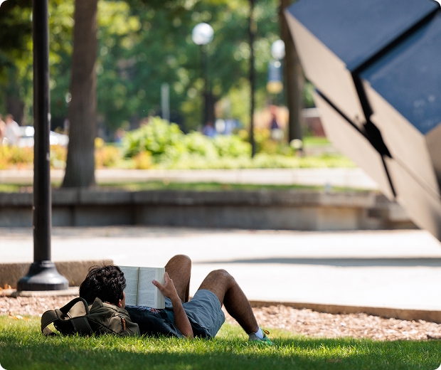 A student lays in the grass reading near the cube